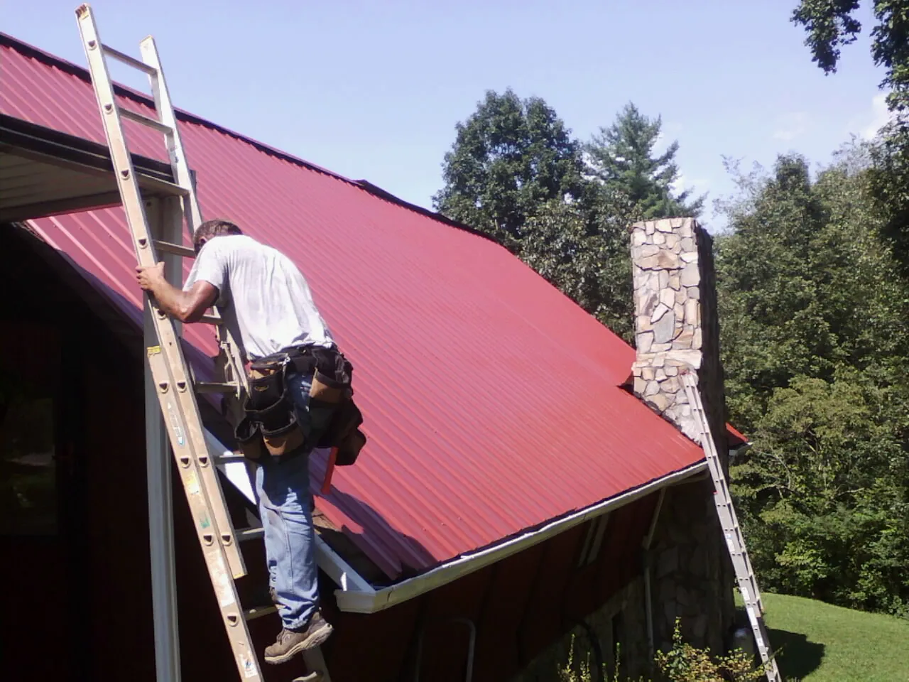 Roofer on a ladder beside a red metal roof and stone chimney — Soffit & Fascia Repair by Roofing Brothers in Gladwyne